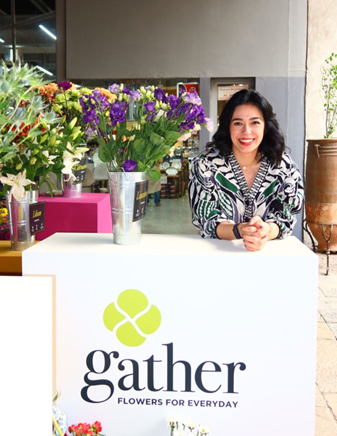 Aida Jorati smiling behind a white branded "gather" counter featuring several buckets of fresh purple and white flowers.